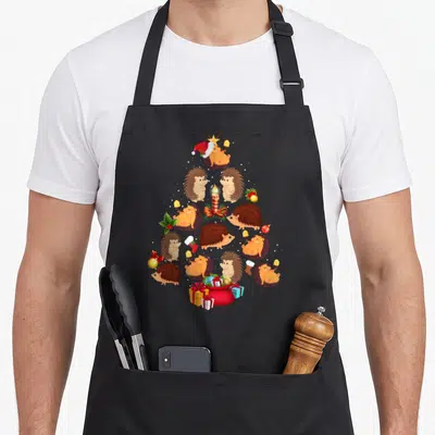 Tablier de cuisine noir avec des hérissons disposés en forme d'arbre de Noël entourés de cadeaux et de décorations festives.