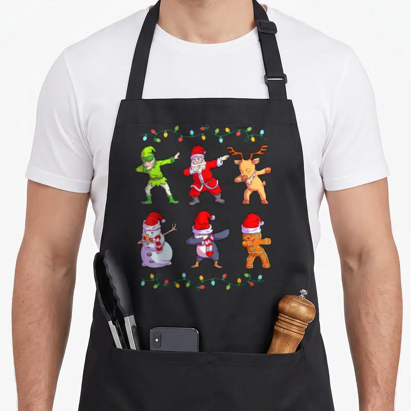 Tablier de cuisine noir avec six personnages de Noël effectuant la danse dab encadrés par des guirlandes lumineuses colorées.
