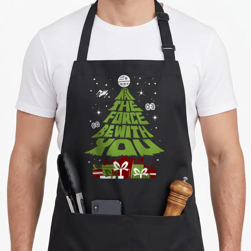Tablier de cuisine noir avec un sapin vert stylisé en texte blanc et des paquets cadeaux rouges et verts au pied.