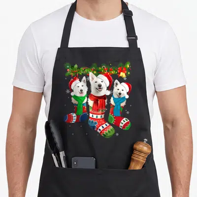 Tablier de cuisine noir avec trois chiens blancs dans des chaussettes de Noël ornées de guimpes et de guirlandes.