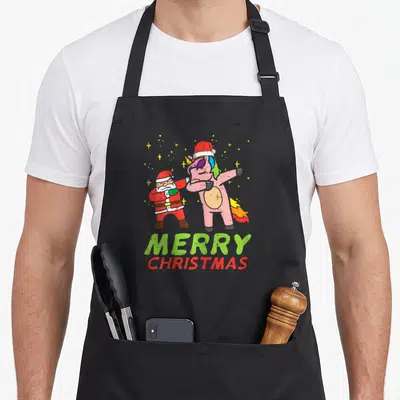 Tablier de cuisine noir avec une licorne rose et le Père Noël effectuant un mouvement de danse sous des étoiles jaunes.