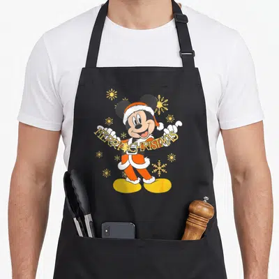 Tablier de cuisine noir avec Mickey Mouse en tenue de Noël tenant l'inscription Merry Christmas Gold parmi des flocons dorés.