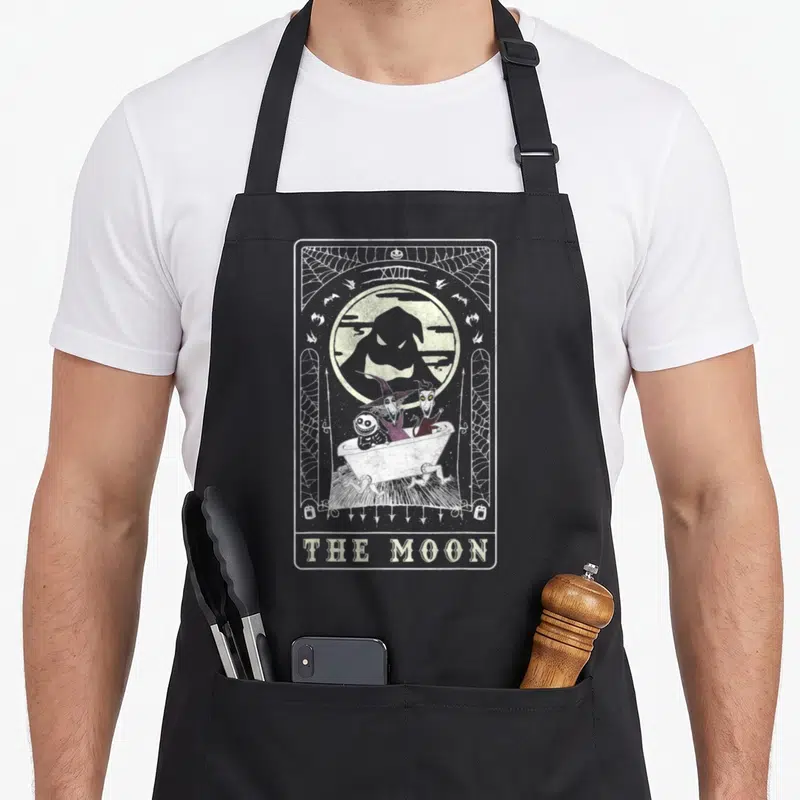 Tablier de cuisine noir avec illustration tarot The Moon montrant Jack, Sally et Zero dans une baignoire sous un visage lunaire.