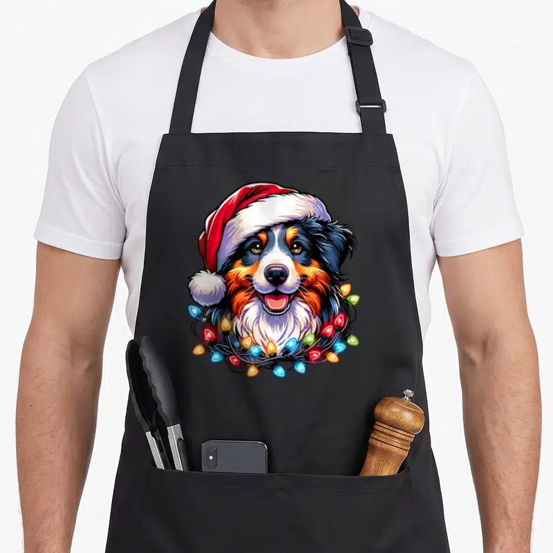 Tablier de cuisine noir avec illustration colorée d'un chien Berger Australien coiffé d'un chapeau de Père Noël et de guirlandes lumineuses.