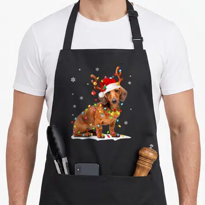 Tablier de cuisine noir avec un teckel portant une casquette de Père Noël et des lumières festives autour du cou.