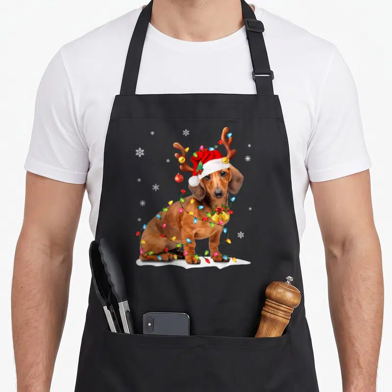 Tablier de cuisine noir avec un teckel portant une casquette de Père Noël et des lumières festives autour du cou.