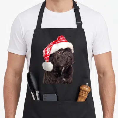 Tablier de cuisine noir avec un bouledogue français portant un chapeau de Père Noël rouge et blanc orné de flocons de neige.