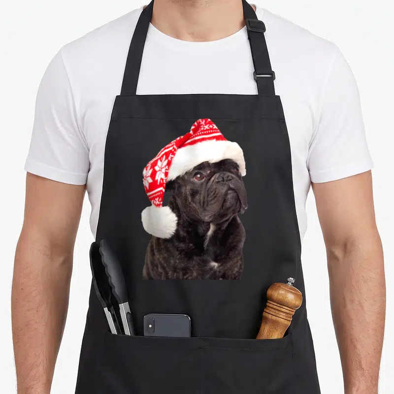 Tablier de cuisine noir avec un bouledogue français portant un chapeau de Père Noël rouge et blanc orné de flocons de neige.