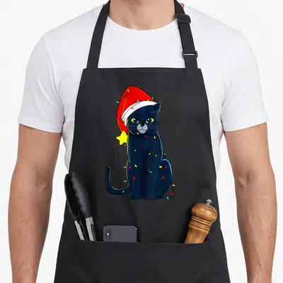 Tablier de cuisine noir avec un chat noir portant un bonnet rouge et des lumières de Noël colorées autour du corps.