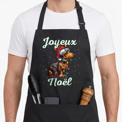 Tablier de cuisine noir avec un teckel portant un bonnet de Père Noël et des guirlandes lumineuses colorées.