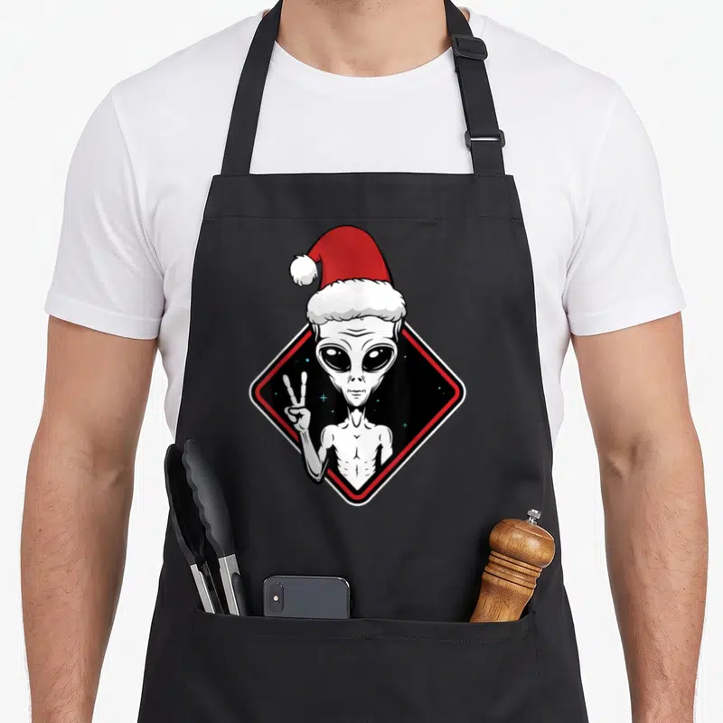 Tablier de cuisine noir avec illustration d'un alien portant un chapeau de Père Noël et faisant un signe de la paix.