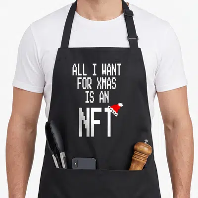Tablier de cuisine noir avec inscription pixelisée blanche All I Want For Xmas Is An NFT et une petite casquette de Père Noël rouge posée sur le T.