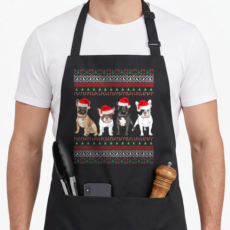 Tablier de cuisine noir avec quatre bouledogues français portant des bonnets de Noël sur un fond à motifs festifs.
