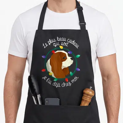 Tablier de cuisine noir avec portrait de chien Cavalier King Charles encadré par des lumières de Noël colorées et un texte blanc.