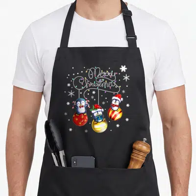 Tablier de cuisine noir avec trois pingouins festifs posés sur des décorations sphériques brillantes et des flocons blancs.