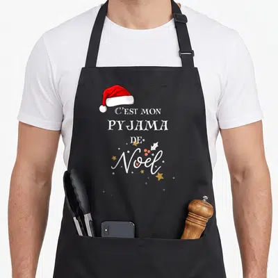 Tablier de cuisine noir avec inscription C'est mon pyjama de Noël et une casquette de Père Noël rouge en haut à gauche.