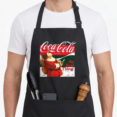 Tablier de cuisine noir avec illustration vintage d'un Père Noël tenant une bouteille Coca-Cola et le texte the Real Thing.