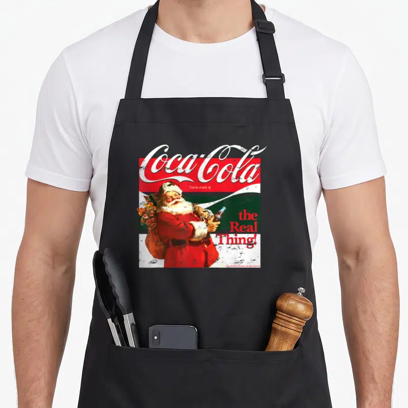 Tablier de cuisine noir avec illustration vintage d'un Père Noël tenant une bouteille Coca-Cola et le texte the Real Thing.