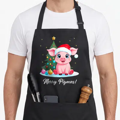 Tablier de cuisine noir avec illustration d'un cochon rose en bonnet de Père Noël devant un sapin décoré.