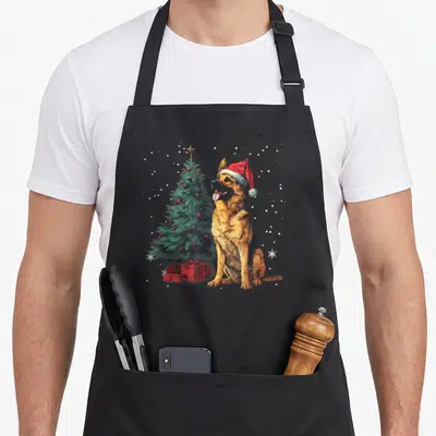 Tablier de cuisine noir avec illustration d'un chien berger allemand portant un bonnet rouge à côté d'un sapin de Noël.