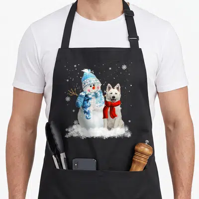 Tablier de cuisine noir avec un Berger Blanc Suisse et un bonhomme de neige en tenue d'hiver sur fond de neige.