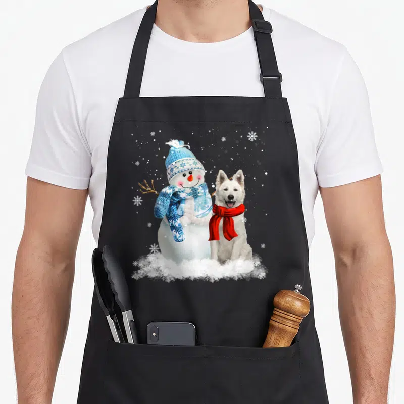 Tablier de cuisine noir avec un Berger Blanc Suisse et un bonhomme de neige en tenue d'hiver sur fond de neige.
