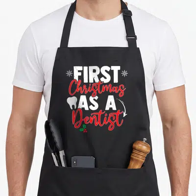 Tablier de cuisine noir avec texte First Christmas As A Dentist en rouge et blanc, orné d'une dent et de flocons.