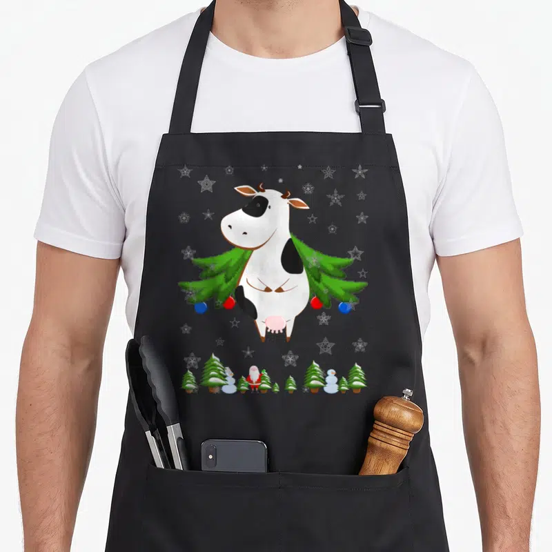 Tablier de cuisine noir avec une vache blanche et noire portant un arbre de Noël décoré de boules colorées.