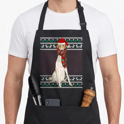 Tablier de cuisine noir avec un Barzoï portant un bonnet de Noël et une écharpe verte et rouge sur fond de motifs hivernaux.