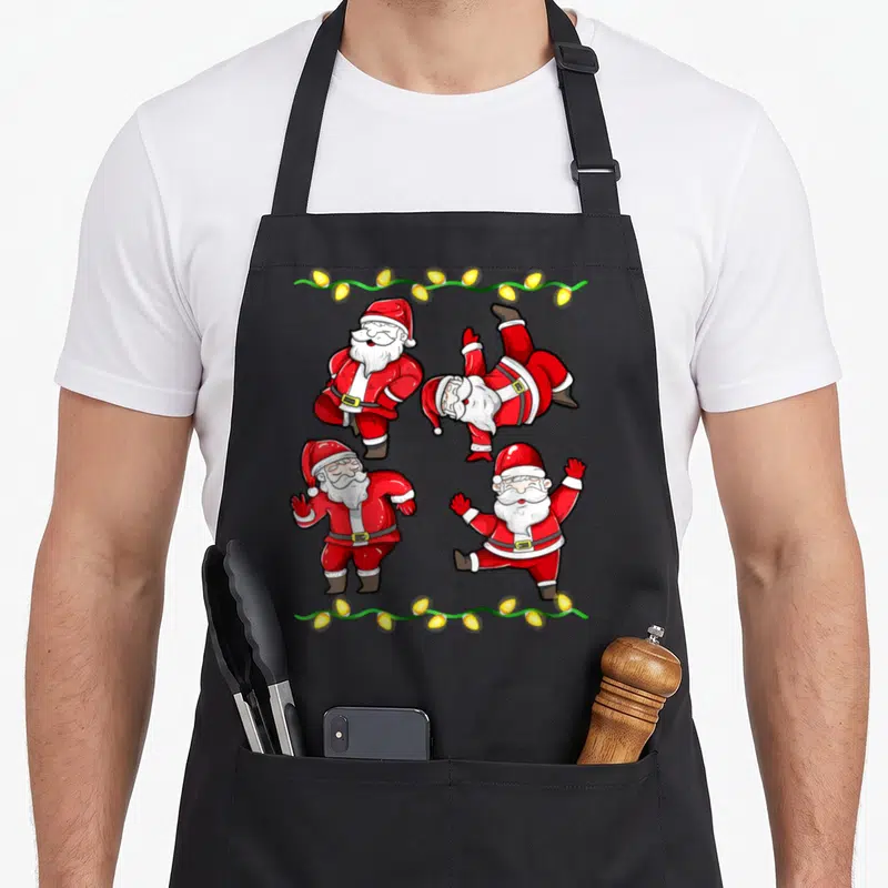 Tablier de cuisine noir avec quatre Pères Noël en train de danser et une guirlande lumineuse jaune sur fond gris.