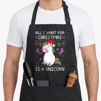 Tablier de cuisine noir avec une licorne blanche au chapeau de Père Noël et texte pixelisé coloré.
