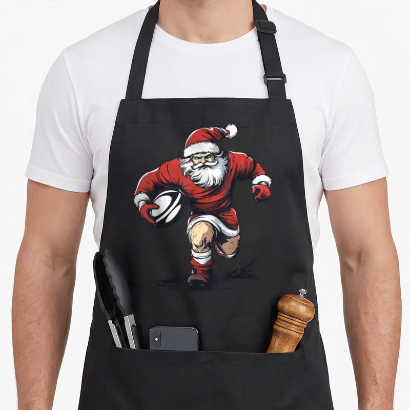 Tablier de cuisine noir avec illustration d'un Père Noël joueur de rugby portant un ballon rouge et blanc.