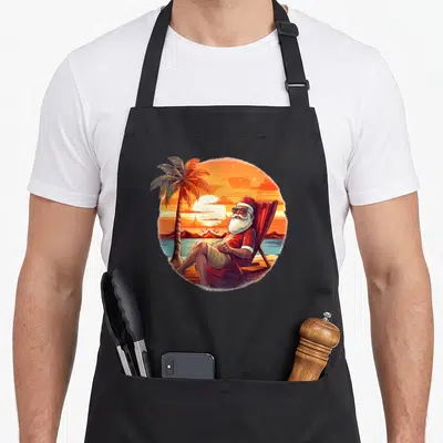 Tablier de cuisine noir avec illustration circulaire d'un Père Noël relaxant sur une chaise bajo un palmier au coucher du soleil.