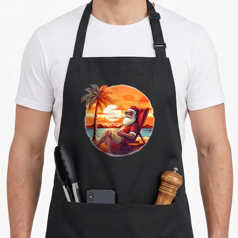 Tablier de cuisine noir avec illustration circulaire d'un Père Noël relaxant sur une chaise bajo un palmier au coucher du soleil.