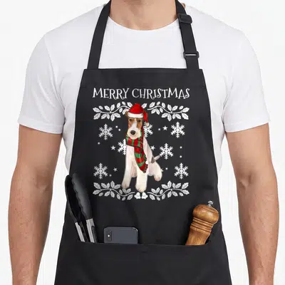 Tablier de cuisine noir avec un Fox Terrier en bonnet rouge et écharpe à carreaux vert et rouge, entouré de flocons et de texte blanc.