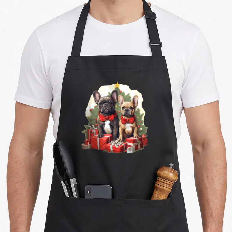Tablier de cuisine noir avec deux bouledogues français en nœuds papillon rouges devant des cadeaux de Noël.