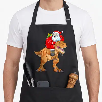 Tablier de cuisine noir avec illustration colorée d'un Père Noël chevauchant un dinosaure T-Rex.