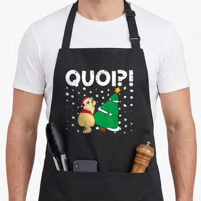 Tablier de cuisine noir avec illustration d'un chat surprise en bonnet de Père Noël à côté d'un sapin de Noël vert.