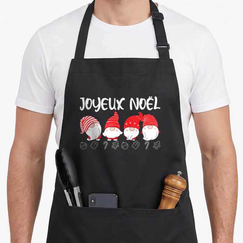 Tablier de cuisine noir avec quatre gnomes de Noël et l'inscription Joyeux Noël au-dessus d'une rangée de décorations.