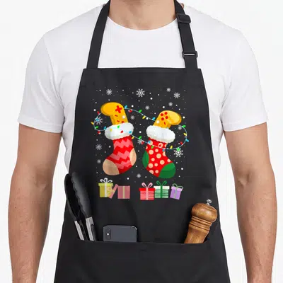 Tablier de cuisine noir avec deux chaussettes de Noël stylisées manettes de jeu et guirlandes lumineuses sur fond neigeux.