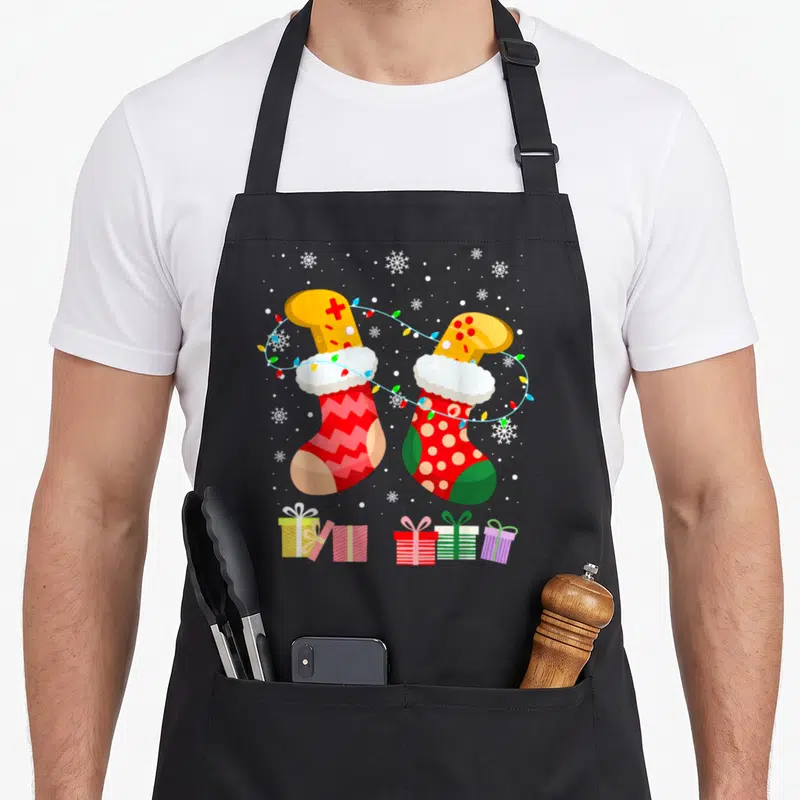 Tablier de cuisine noir avec deux chaussettes de Noël stylisées manettes de jeu et guirlandes lumineuses sur fond neigeux.