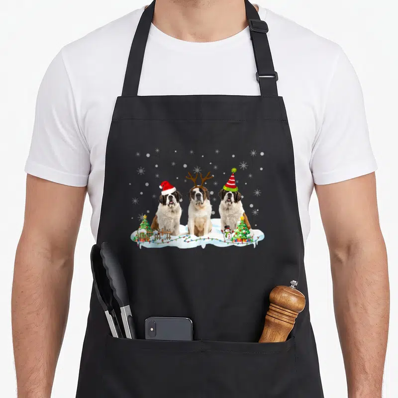 Tablier de cuisine noir avec trois chiens Saint Bernard déguisés assis dans la neige près d'arbres de Noël et de guirlandes lumineuses.