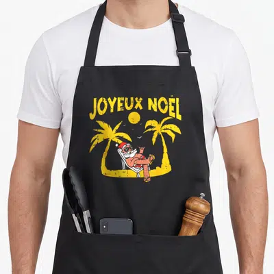 Tablier de cuisine noir avec illustration d'un Père Noël relaxant sur une chaise de plage entre deux palmiers jaunes.