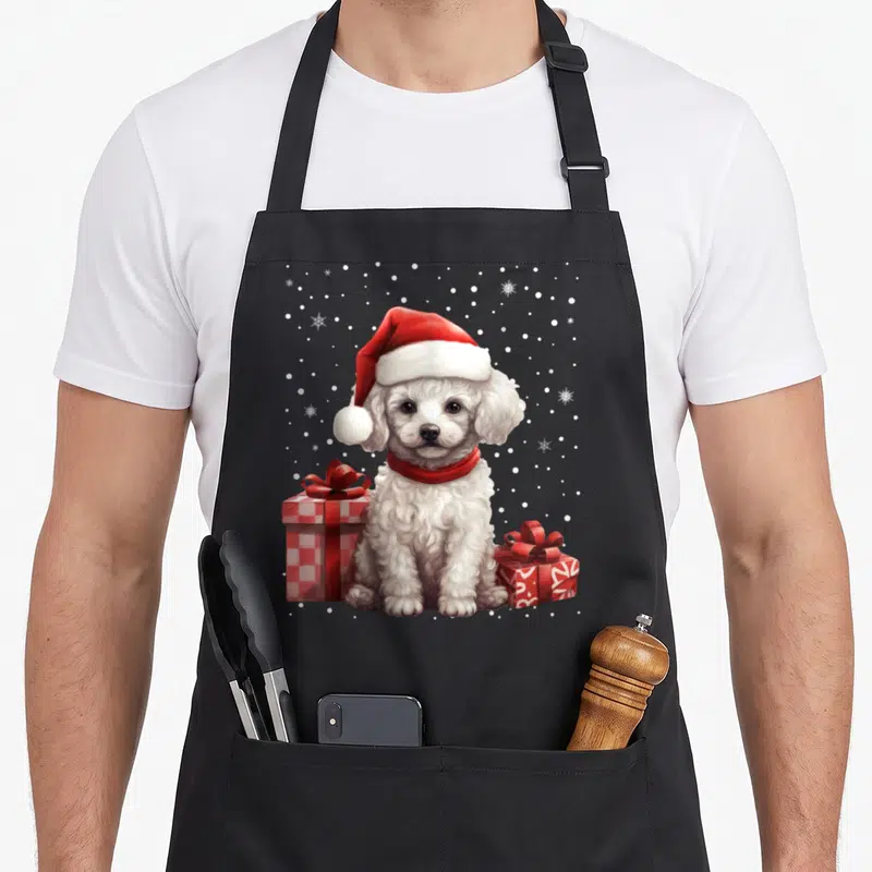 Tablier de cuisine noir avec un chiot caniche blanc coiffé d'un bonnet rouge et des cadeaux empaquetés.