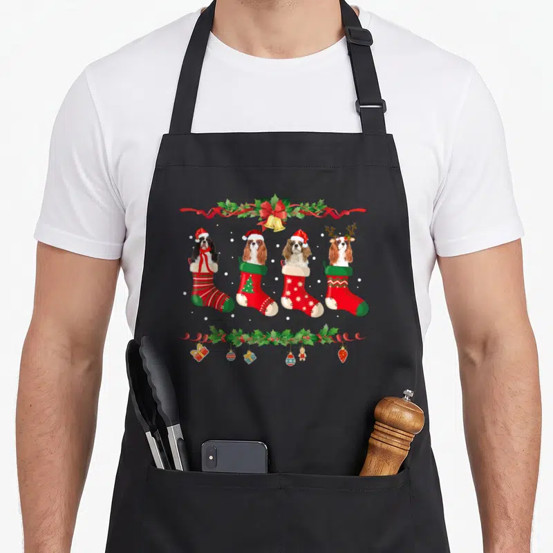 Tablier de cuisine noir avec quatre chaussettes de Noël contenant des chiens Cavaliers King Charles Spaniel et des décorations festives.