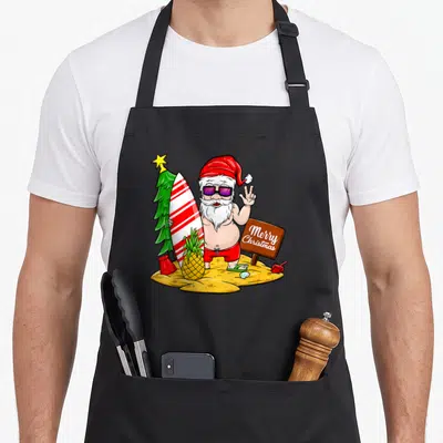 Tablier de cuisine noir avec illustration colorée d'un Père Noël en maillot de bain tenant une planche de surf à côté d'un panneau Merry Christmas.