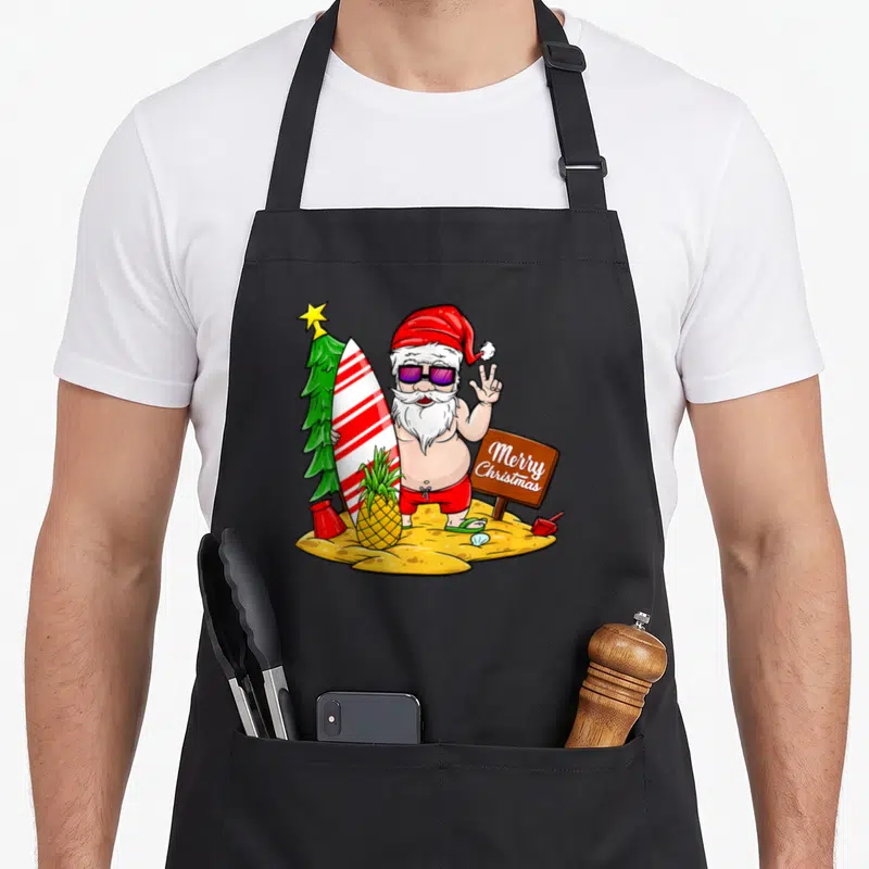 Tablier de cuisine noir avec illustration colorée d'un Père Noël en maillot de bain tenant une planche de surf à côté d'un panneau Merry Christmas.
