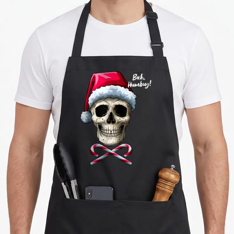 Tablier de cuisine noir avec un crâne portant une toque rouge et blanche surmontée de deux cannes à bonbons croisées.