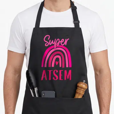 Tablier de cuisine noir avec arc-en-ciel rose vif, cœur central et texte Super ATSEM en lettres stylisées.