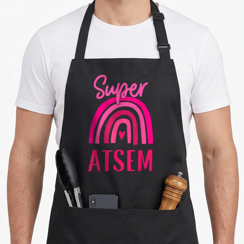 Tablier de cuisine noir avec arc-en-ciel rose vif, cœur central et texte Super ATSEM en lettres stylisées.